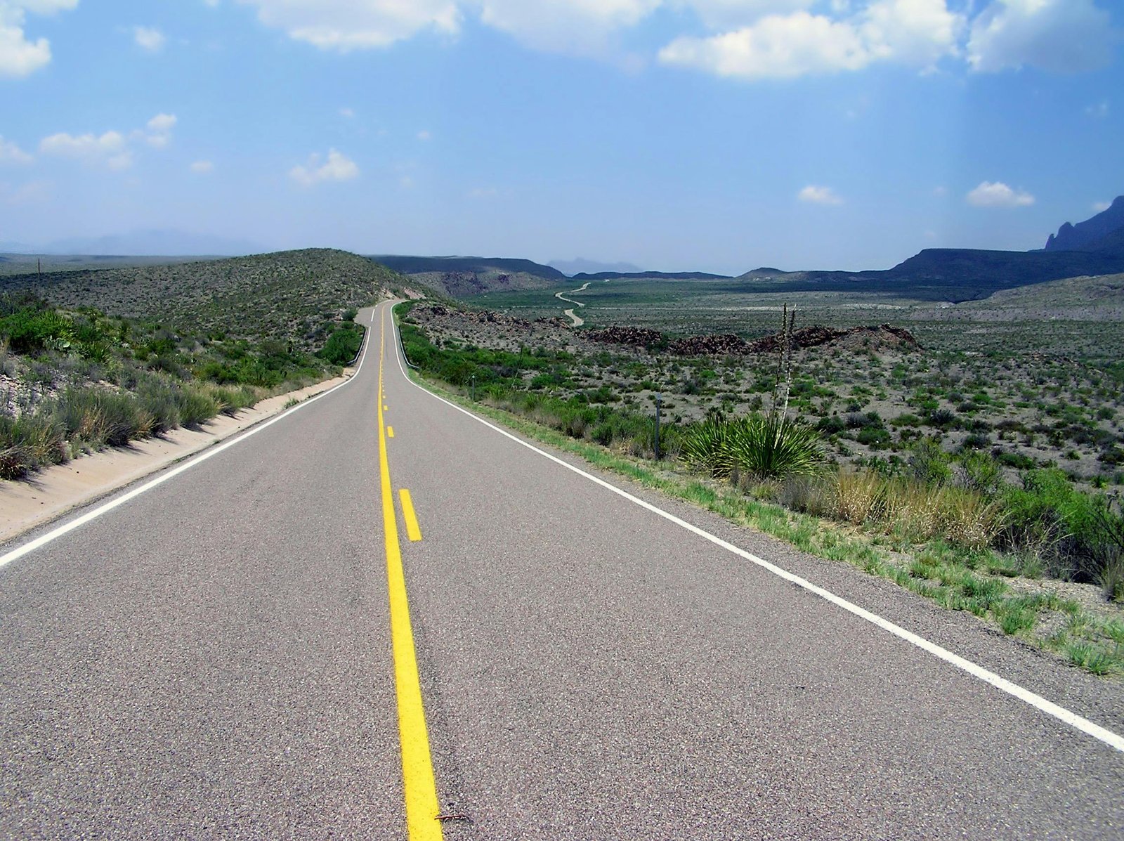 A long open road stretches through a scenic desert landscape under a bright blue sky.
